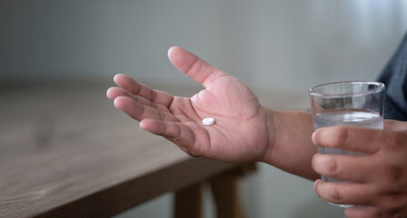 man hand holds a white pill or capsule and a glass of water at home, eating vitamin on time,...