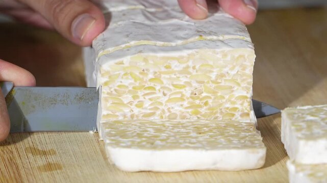 Closeup slicing tempeh block on wooden board. Fermented soybean texture appears clean, dense, natural
