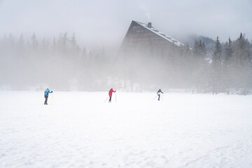 Cross-country skiers glide across a frozen, snow-covered lake during a misty winter day, featuring a modern A-frame mountain hotel and pine forest in the heavy white fog.