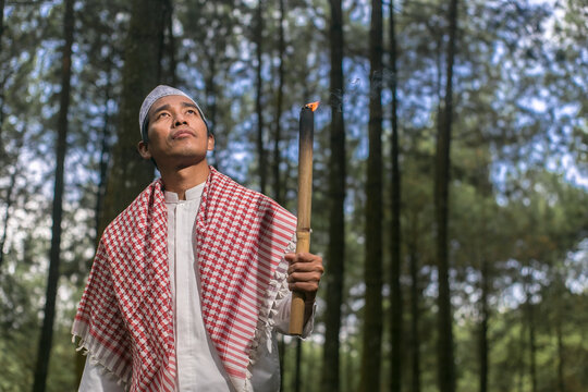 An Asian Indonesian man stands holding a torch wearing Muslim attire, a peci and sarong against a natural background