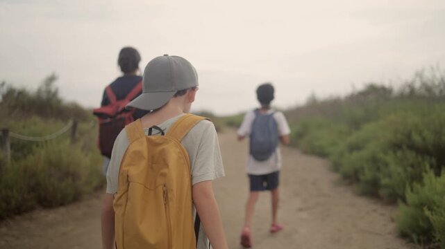 Family walking on a wooden boardwalk, mother and two children with backpacks exploring sandy dunes, enjoying outdoor summer travel, holiday, and togetherness on a beach path