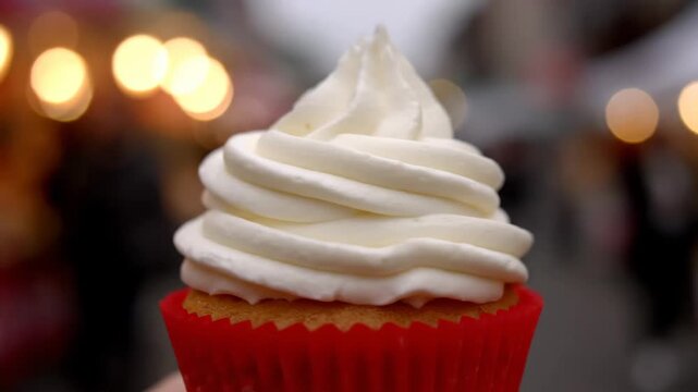 Delicious close-up of a vanilla cupcake with thick white piped cream frosting in a vibrant red wrapper against a warm festive bokeh background.