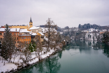 Obraz premium The picturesque town of Novo Mesto in Slovenia during winter, covered in snow, showing beautiful facades and houses along the colorful Krka River. Novo Mesto, Slovenia 09.01.2026 