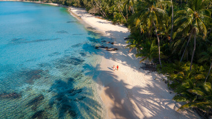 Naklejka premium A couple experiences the tranquility of Takhian Beach on Koh Kood, Thailand, where two visitors stroll along the soft, powdery sands beneath tropical palm trees, embraced by the azure sea.