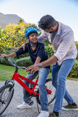 Fototapeta premium Asian father and son balancing on bicycle at garden patio wearing blue helmet, watch and sneaker