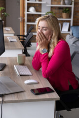 Office worker covering mouth while yawning at modern office desk with laptop, keyboard, coffee mug