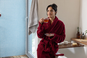 Woman wearing bathrobe sipping tea from glass teacup on bathtub edge in bathroom with bottles