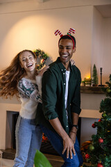 Naklejka premium Diverse couple dancing playfully in festive living room with reindeer headband, christmas tree