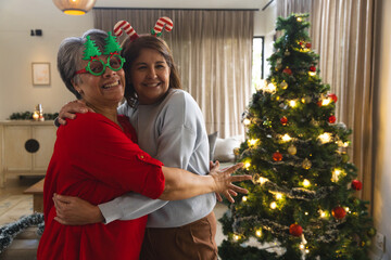 Obraz premium Senior mother and daughter hugging in living room by Christmas tree wearing candy-cane headband