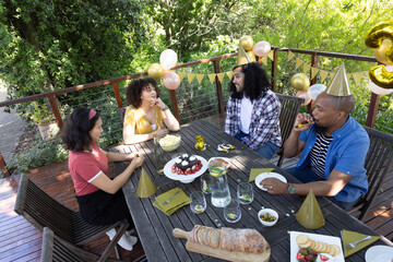 Diverse friends gathering around backyard deck table with berry-topped cake, gold party hats