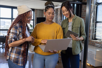 Diverse female coworkers collaborating around laptop and tablet in coworking office with whiteboard