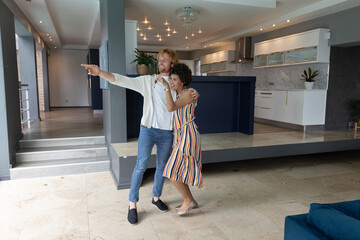 Naklejka premium Diverse couple standing in open-plan home area, embracing while pointing at kitchen island
