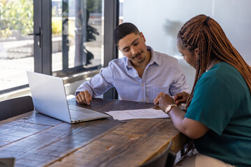 Diverse coworkers reviewing printed document on wooden table in meeting room with laptop