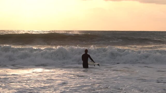 Surfer in warm wetsuit enter water with board at sunset. Winter sports in Italy. Health, sea, and nature.