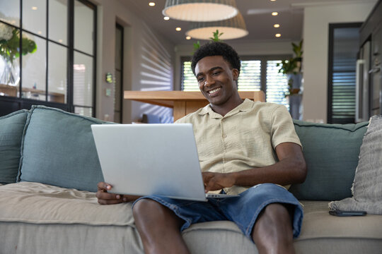 African american man sitting on sofa using laptop with smartphone in living room - Powered by Adobe