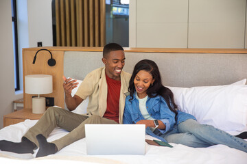 African American couple sitting on bed using laptop in modern bedroom with lamp and smartphone