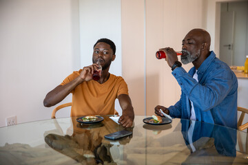 African american father, son sipping red juice, eating avocado toast in kitchen with smartphone