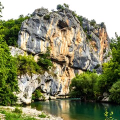 A scenic view of a rocky cliff face beside a river, surrounded by lush green trees on a sunny day