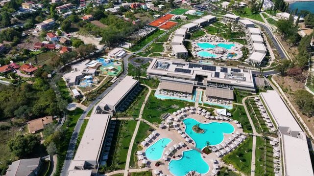 High-altitude aerial drone view of the expansive Ikos Odisia luxury resort in Corfu, Greece. Flying over the hotel, tennis courts, and swimming pools on the Ionian coast.