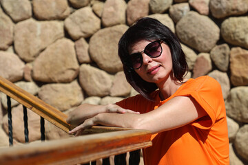 Elegant woman in orange dress standing against rustic stone wall, wearing outdoors.