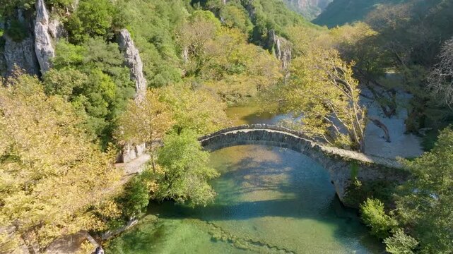 Aerial drone pullback shot of the historic Klidonia stone bridge over the Voidomatis River, revealing a weir and clear water in the Vikos-Aoos Geopark, Zagori, Greece