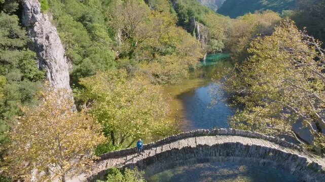 Aerial drone pulls back to reveal a person on the historic Klidonia stone bridge, spanning the clear Voidomatis River in the Zagori region of Epirus, Greece