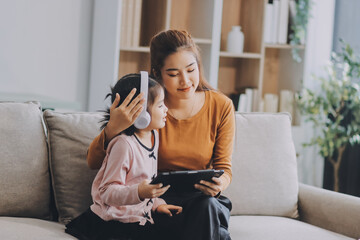 Obraz premium A cheerful Asian mother helps her young daughter do homework and color a book at home, while the father works on a laptop in the background. A cozy, bright, and modern family lifestyle.