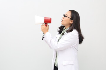 Fototapeta premium Asian adult female doctor wearing doctor's coat, stethoscope hanging around neck, white studio background, carrying and speaking through megaphone, delivering announcement, health information concept.