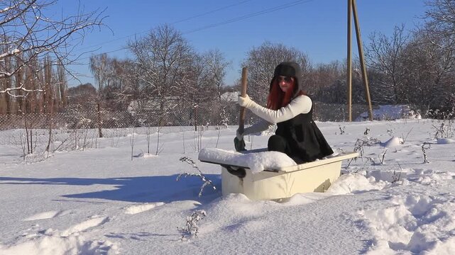 A woman with red hair in winter clothes sits in a snowy bathtub in the yard and holds a shovel, a humorous scene on a frosty sunny day.