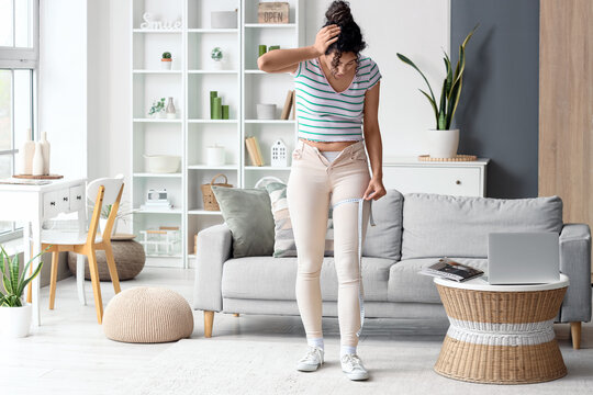 Shocked African-American woman in tight pants measuring her thigh at home. Diet concept