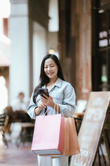Happy Asian woman holding shopping bags outdoors in a trendy urban area, smiling confidently after shopping, representing modern lifestyle, retail experience, and consumer happiness.