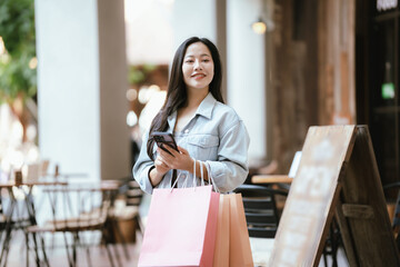 Happy Asian woman holding shopping bags outdoors in a trendy urban area, smiling confidently after shopping, representing modern lifestyle, retail experience, and consumer happiness.