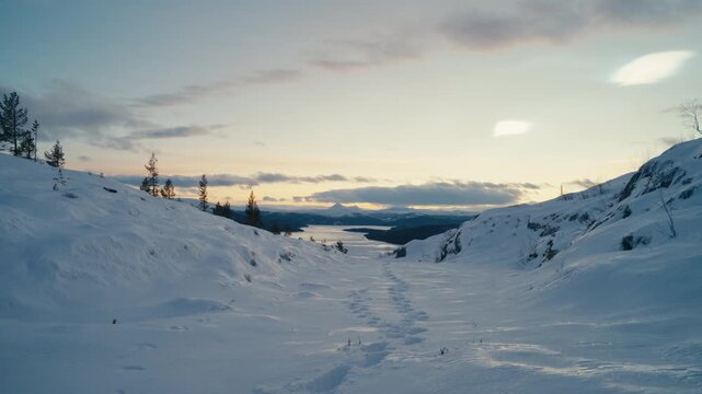 Snow-covered trail winds through quiet mountain terrain at sunset, with deep footprints leading toward a distant frozen lake.