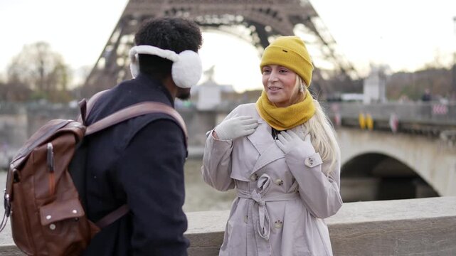 Young diverse couple dressed in winter clothes having a lively conversation on a bridge over the seine river with the iconic eiffel tower in the background, enjoying a romantic trip in paris