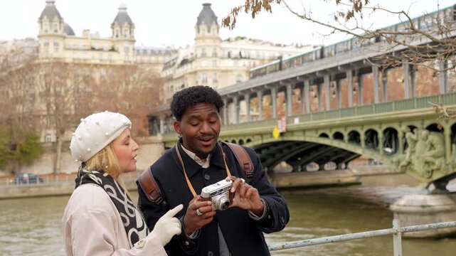 Young multiracial couple visiting paris, france, enjoying a romantic getaway while taking photos with a vintage analog camera with the bir hakeim bridge and the seine river in the background