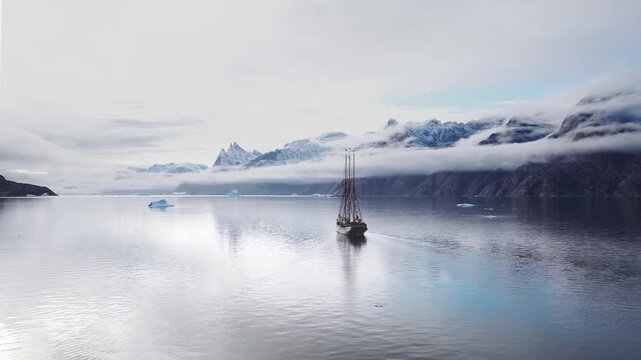 A schooner sails through a Greenlandic fjord with misty snow covered mountains in the background.