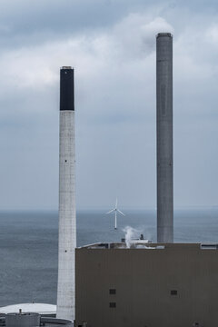 Industrial power plant smokestack chimney with wind turbine energy creates contrast in sky over modern infrastructure showing transition landscape
