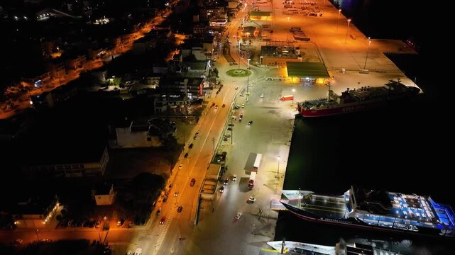 Night aerial drone view of the Port of Igoumenitsa, Greece. Flying over the busy ferry terminal with docked ships, trucks, and car traffic on the illuminated coastal road