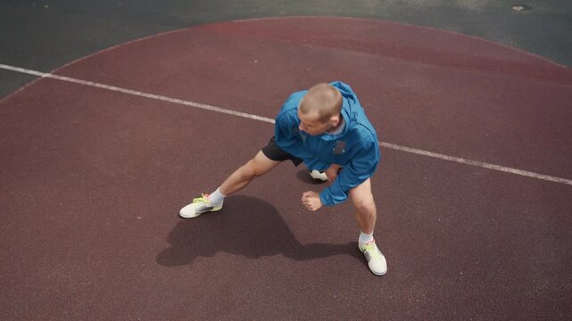 Caucasian man on wet court dribbling in low wide stance, street trainer executing lateral lunges and feints behind ball in blue jacket and white trainers reflective rubber surface, tactical solo