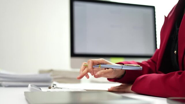 Close up of a female office worker writing on a clipboard and using a computer at her desk, with a stack of documents nearby in a professional, minimalist and modern corporate environment