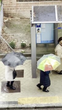 Paraguas y abrigo con lluvia, tormenta y nubes de agua con pareja andando por acera de calle mojada con parada de bus