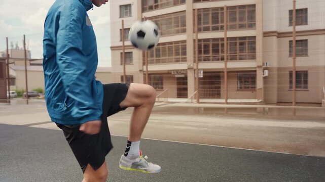 young man juggling football on wet asphalt, urban street under light rain, blue jacket, black shorts, white sneakers, puddles splashing, closeup leg control, midair ball, walking away shots,