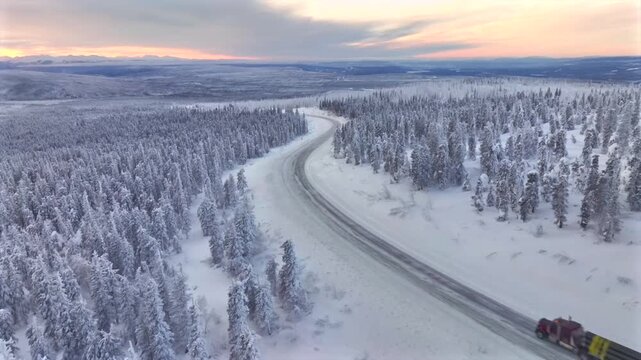 Aerial view of a red truck driving on the Dalton Highway, surrounded by snow-covered trees under a pastel sky, Alaska, United States.