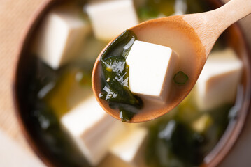 Close up of wooden spoon scooping miso soup with soft tofu and seaweed from a wooden bowl, Top view