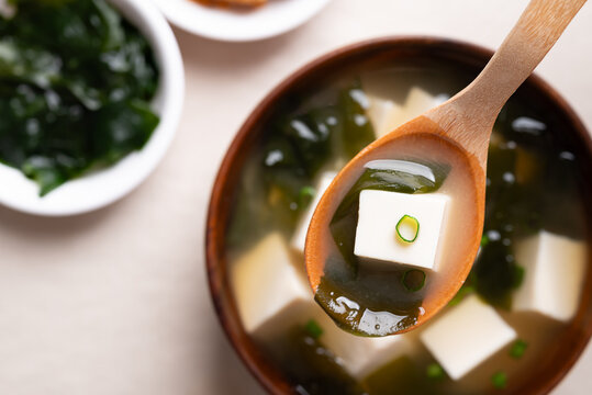 Close up of wooden spoon scooping miso soup with soft tofu and seaweed from a wooden bowl, Top view