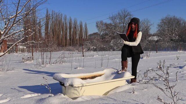 A beautiful girl with a diary in her hands near an old bathtub in the middle of a snowy landscape plans her day. A combination of fashion, nature and an old bathtub.