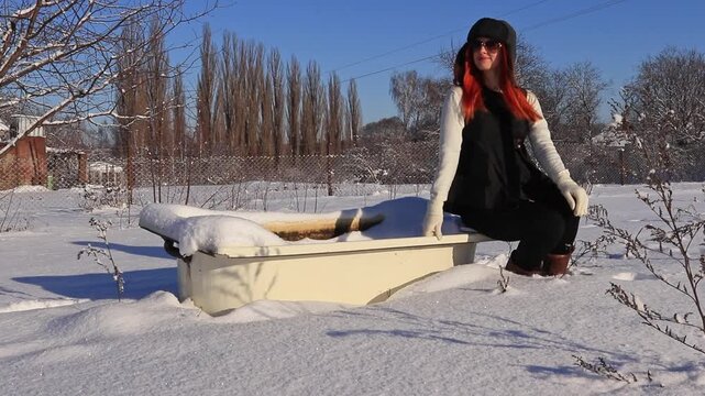 Winter lifestyle shot: a stylish redhead woman in warm clothes sits by a snowy bathtub in the yard, creating an atmosphere of calm, cold and creative mood.
