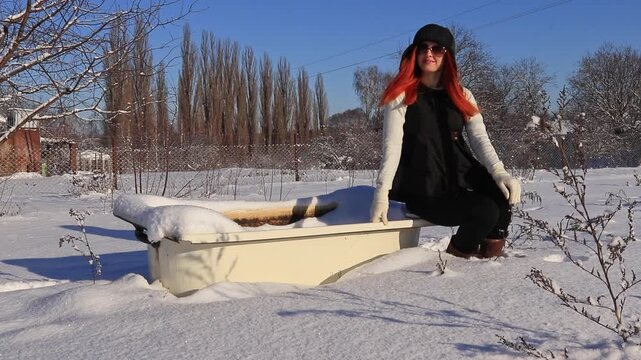 Stylish woman with red hair poses near an old bathtub in a snowy landscape. A combination of fashion, nature and an old bathtub. Winter fashion mood, contrast of style and cold.