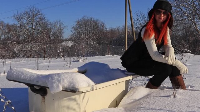 Winter lifestyle shot: a stylish redhead woman in warm clothes sits by a snowy bathtub in the yard, creating an atmosphere of calm, cold and creative mood.