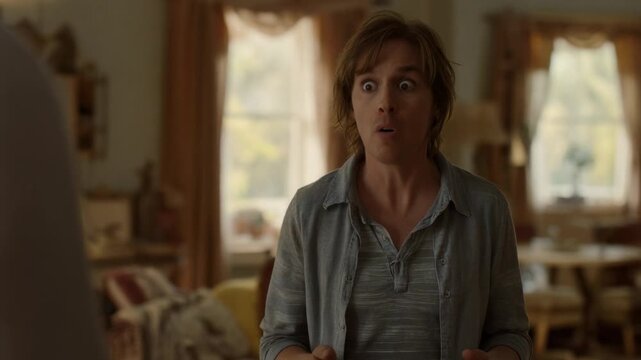 Reacting to offframe remark, man widening eyes, shouting by sofa in living room wearing gray henley